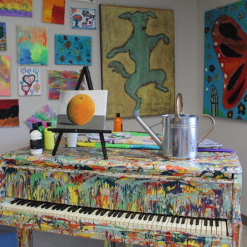 A paint splattered piano with a painting of an orange on a table easel and paint supplies. The piano sits in a corner, and on the walls are brightly colored works of art.