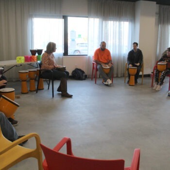 People sit in colorful chairs with bongo drums as they perform in a drumming circle. They are inside a big room with grey floor, large windows.