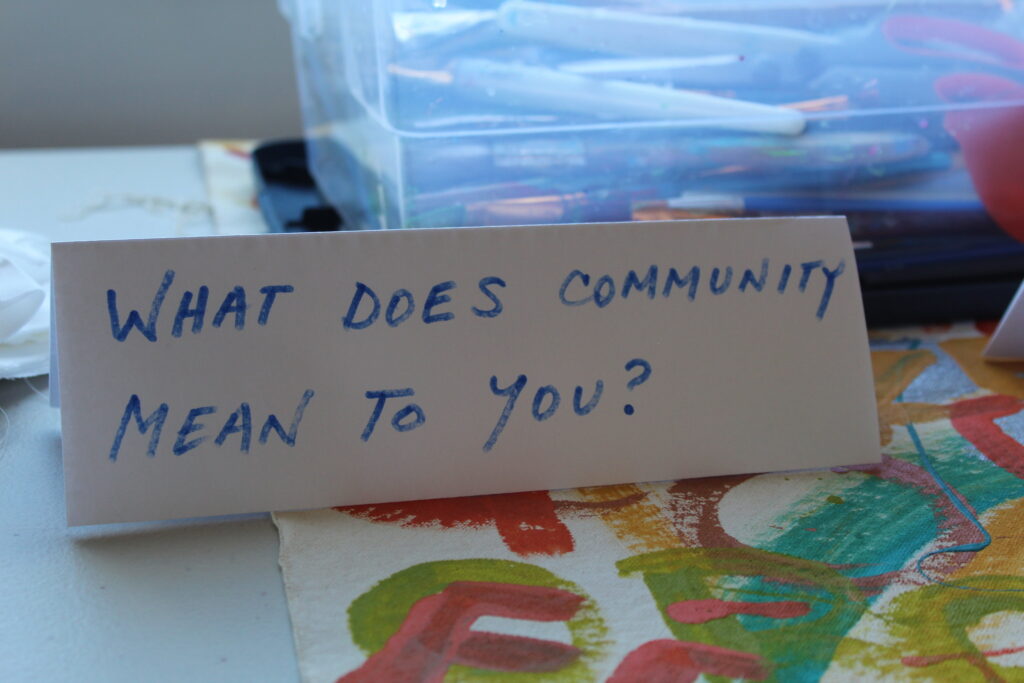 A sign that says, "What does community mean to you?" is placed on a table with art supplies and art work.