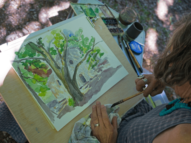A woman sits outside and looks down at her plein air painting of a landscape on her lap. Her brush is in her hand and she has a palette of colors to her right.