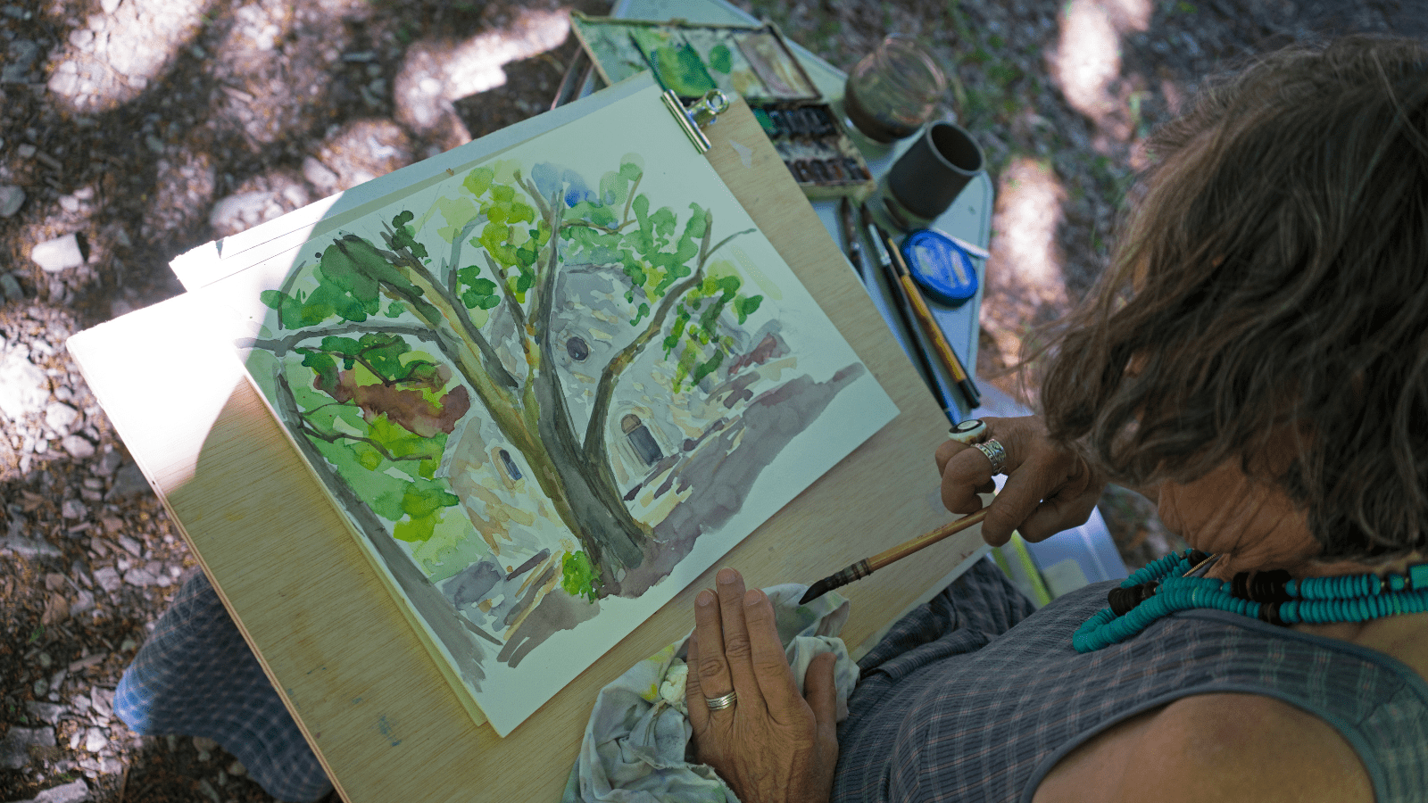 A woman sits outside and looks down at her plein air painting of a landscape on her lap. Her brush is in her hand and she has a palette of colors to her right.