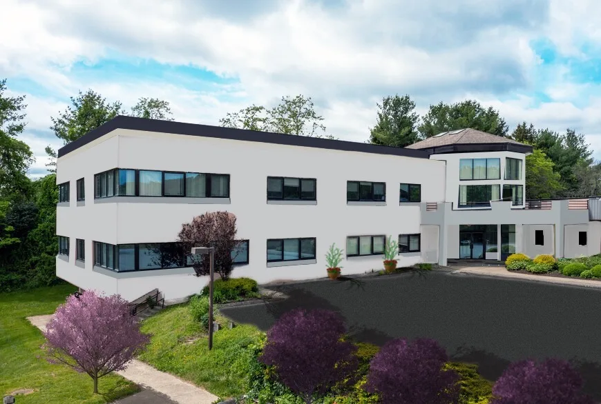 The exterior of the Hudson Valley InterArts Center, a large soft white industrial building with grey trim and black windows, surrounded by trees and a bright sky.