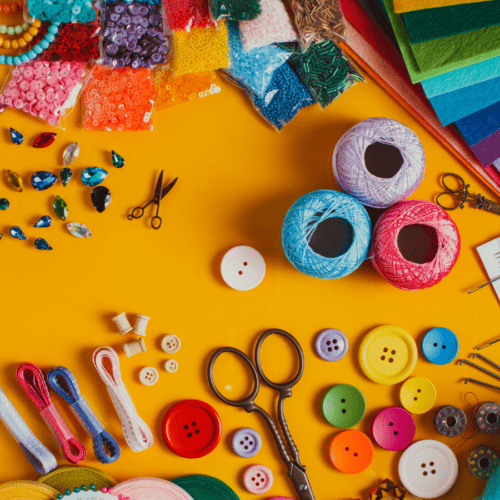 A variety of arts and crafts materials such as yarn, buttons, jewels, scissors, colorful papers are arranged on a bright yellow-orange table top.