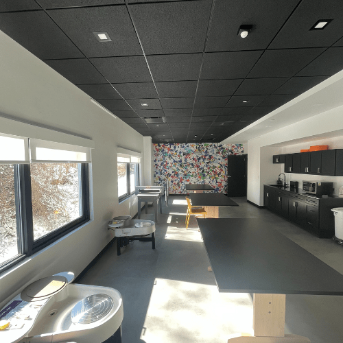 A Ceramics studio with two potters wheels, three black topped tables, a slip basin, cabinets and sink and shelving. The room has a grey floor, white walls, large windows throughout and a black ceiling. There is a bright splash painted wall covering on one of the walls.