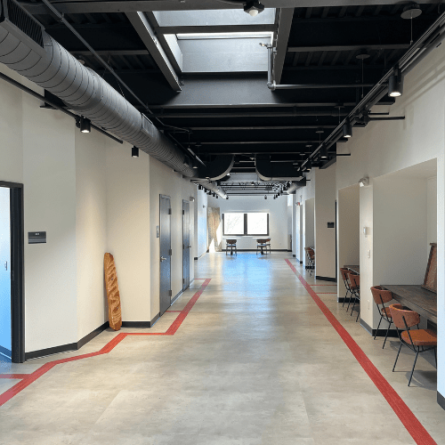 A long hallway with built in desks and chairs for co-working under a black industrial ceiling. At the distant end of the hall are two round cafe tables with a coffee station.
