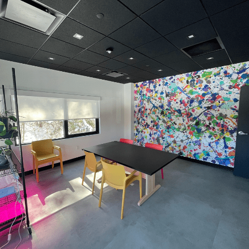 Fiber arts studio with a table, colorful chairs, a splash painted wall covering, shelving and large windows. Grey floor, white walls, black ceiling.
