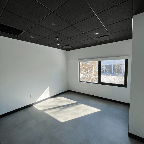 Healing arts studio with grey vinyl flooring, white walls, large windows with light streaming through and black open ceiling.
