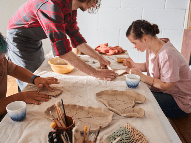Two men and a woman work on clay projects around a table. They work with their hands and simple tools. Two clay pieces, shaped like giant leaves sit on the table.
