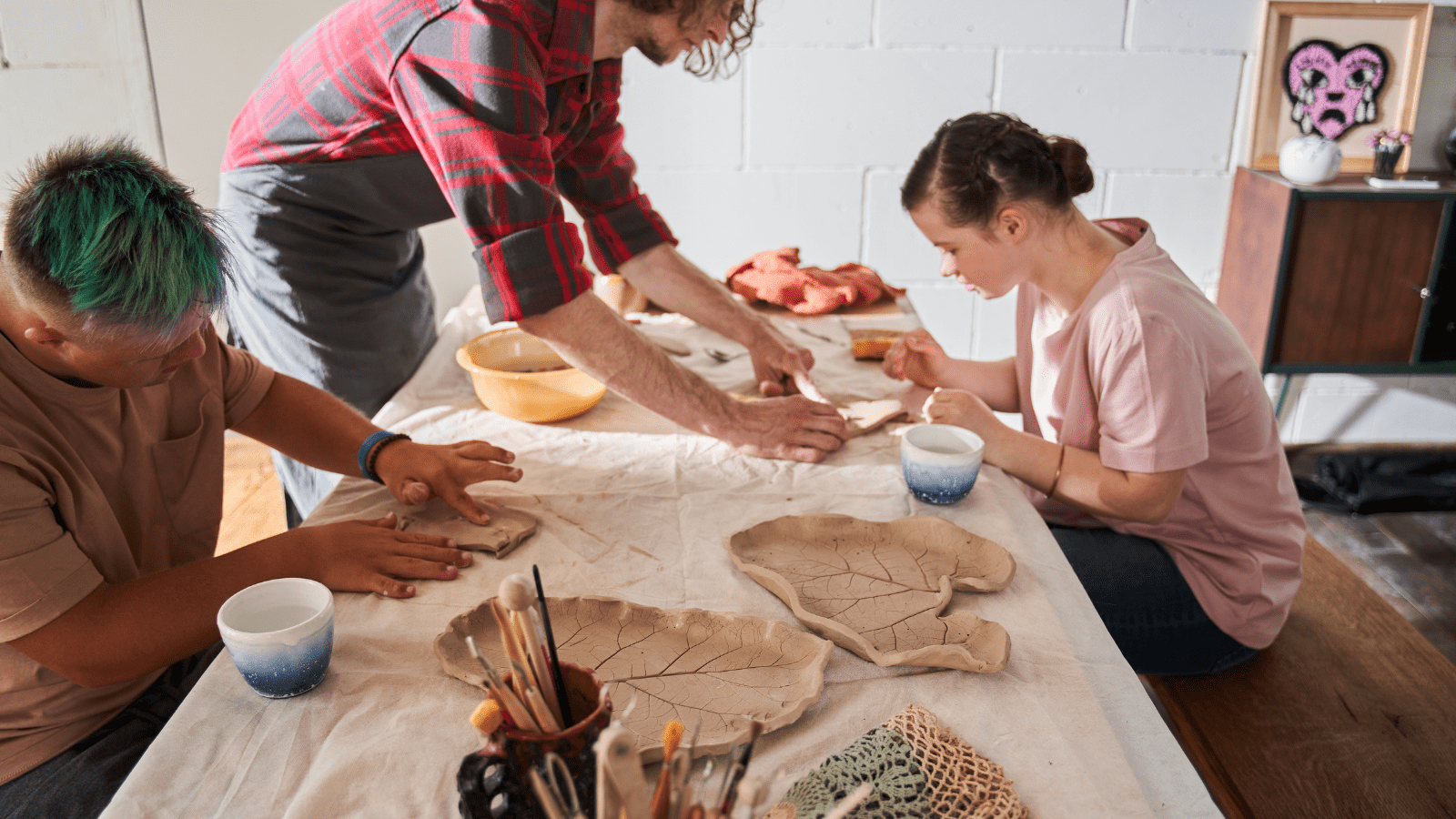 Two men and a woman work on clay projects around a table. They work with their hands and simple tools. Two clay pieces, shaped like giant leaves sit on the table.