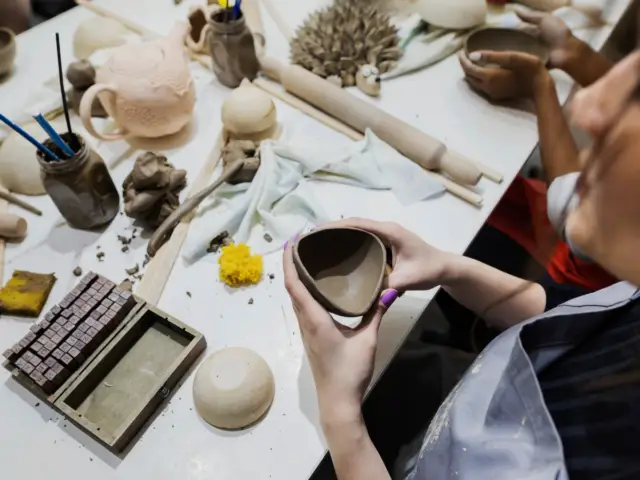 A woman sits around a table in a ceramics class. On the table are various tools used in working with pottery and she holds an unfired triangular pot in her hands.