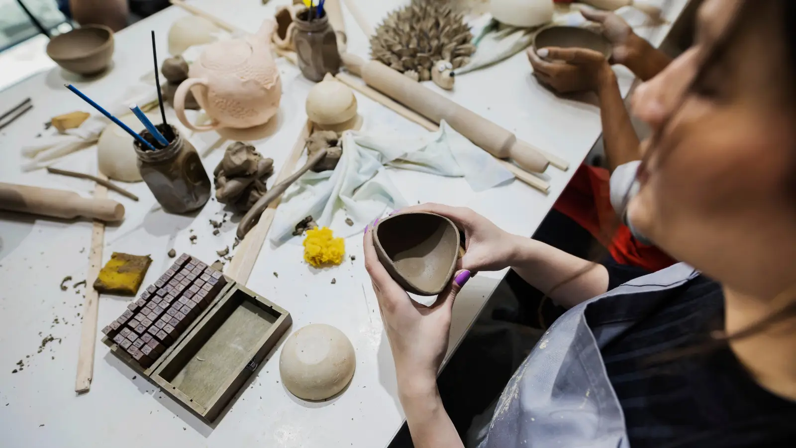A woman sits around a table in a ceramics class. On the table are various tools used in working with pottery and she holds an unfired triangular pot in her hands.
