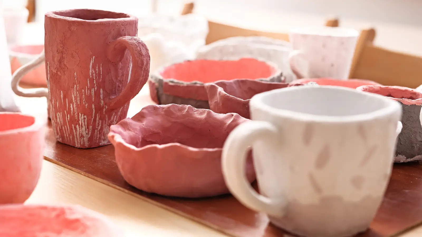 Hand made ceramic mugs and pinch pots glazed in warm reds, oranges and white sit on a wooden table.