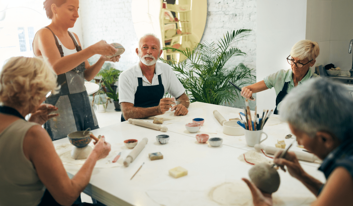 A group of adults sit around a white table working on their clay pots. Some are forming them and others are painting their pots. There are tools and colored glazes on the tables. The room is light filled with a green fern plant in the corner.