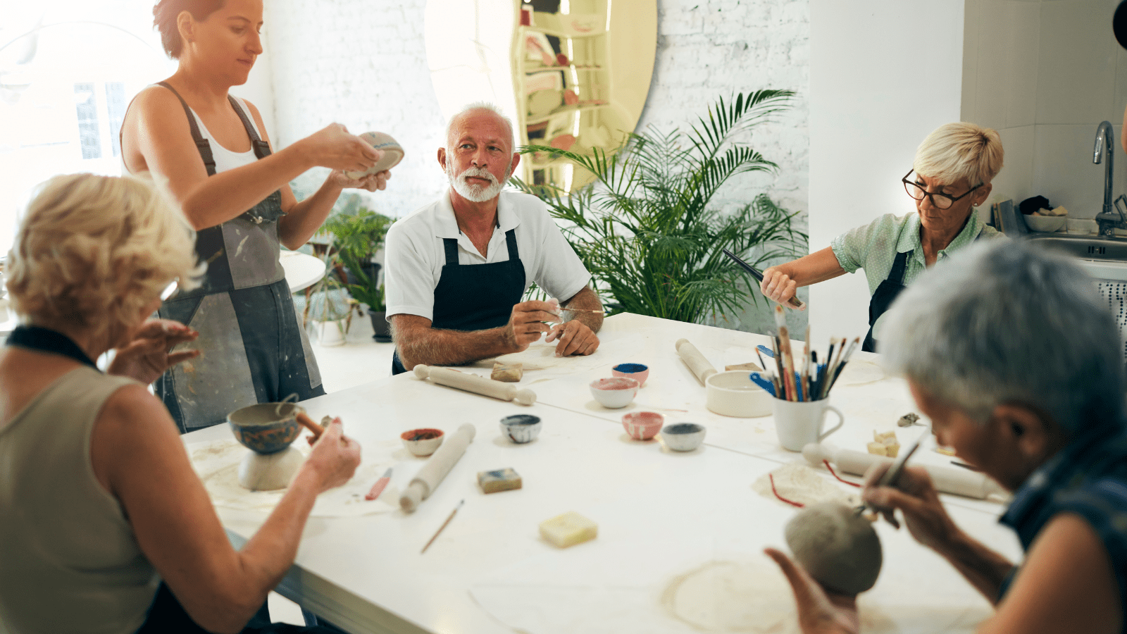 A group of adults sit around a white table working on their clay pots. Some are forming them and others are painting their pots. There are tools and colored glazes on the tables. The room is light filled with a green fern plant in the corner.