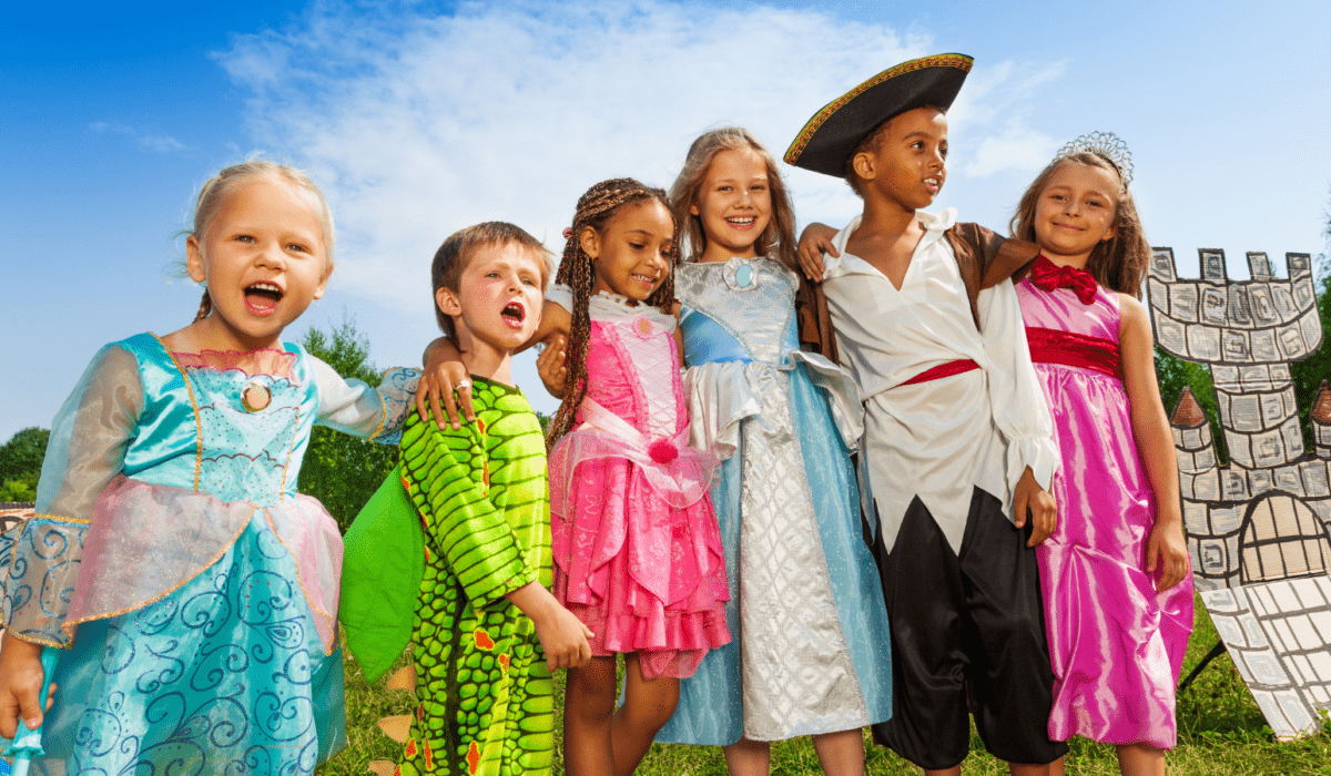 Children in costumes depicting princesses, a pirate and dragon stand in a line outdoors with a hand made scene of a castle in the background.