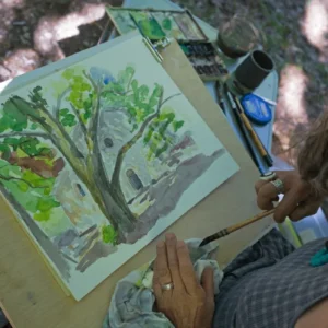 an painter wipes their paintbrush while looking at their landscape painting of a tree in a forest.