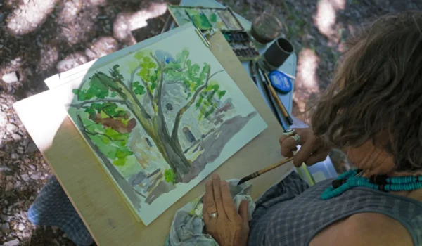 an painter wipes their paintbrush while looking at their landscape painting of a tree in a forest.