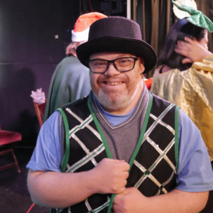 A man with glasses and a short beard wears a theater costume consisting of a bowler hat and a festive vest. He stands back stage and other performers are in the background.