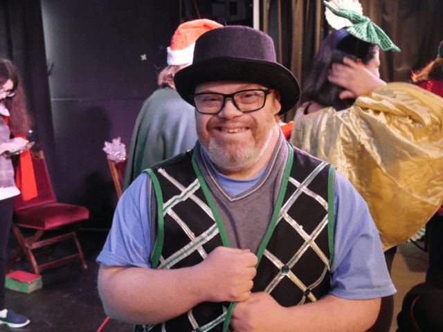A man with glasses and a short beard wears a theater costume consisting of a bowler hat and a festive vest. He stands back stage and other performers are in the background.