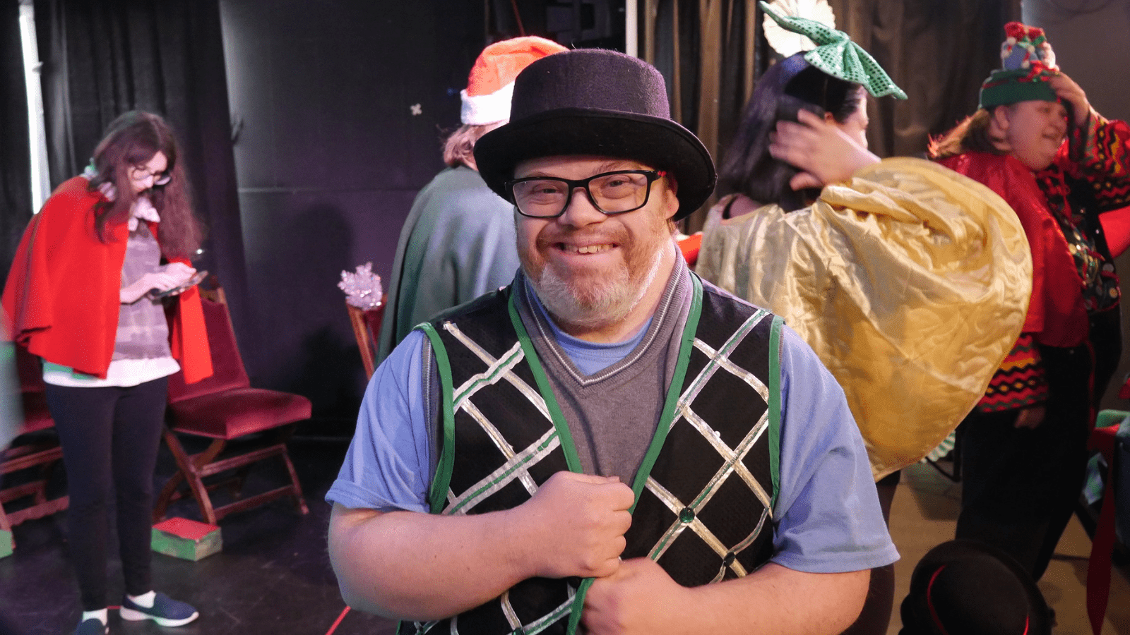 A man with glasses and a short beard wears a theater costume consisting of a bowler hat and a festive vest. He stands back stage and other performers are in the background.