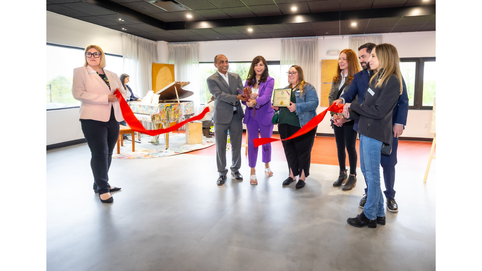 Chief Strategy Officer Bhakar Singh stands with Chief Executive Officer Vicki Sylvester PH.D. as they cut a red ribbon with a giant pair of scissors. The ribbon is held by Paula Hernandez of the Brewster Chamber of Commerce and Congressman Mike Lawler. They stand in the great room of the Hudson Valley InterArts Center and in the background is a piano decorated in splash paint style.