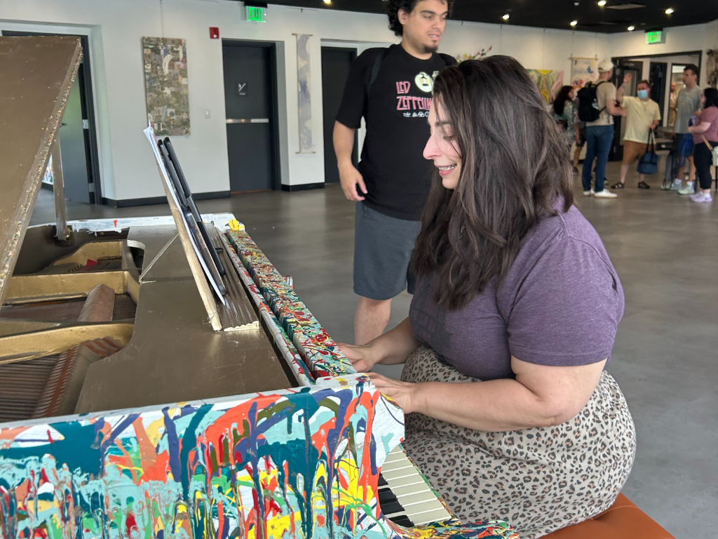 Krista Libertino sits at a piano that has been splash painted. She plays music for the class.